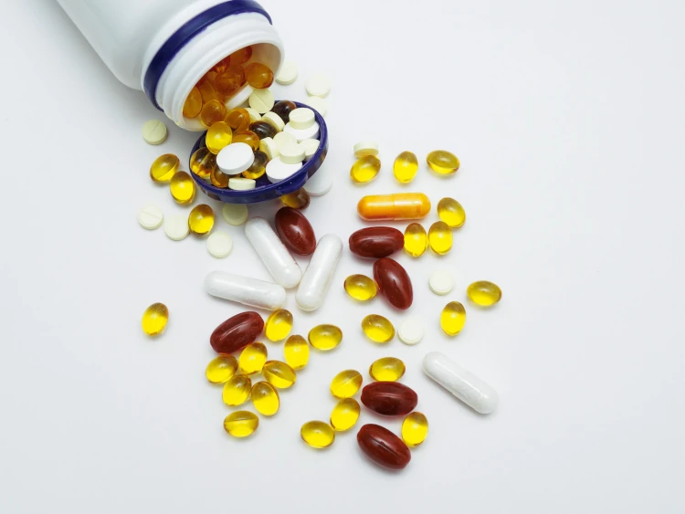 Different vitamins and tablets in capsules of different colors are poured from a white plastic jar on a white background.