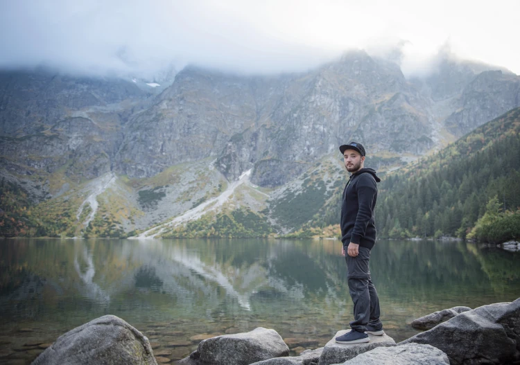 A man in a trucker hat standing near a mountain lake.